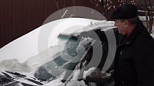 A man cleans a car windshield from snow with a brush. Winter in Russia, the car was covered with snow