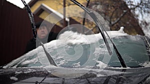 A man cleans a car windshield from snow with a brush. Winter in Russia, the car was covered with snow