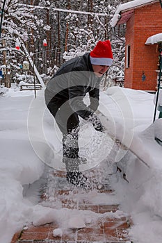 A man cleans steps from snow. In a red hat of Santa Claus and a leather black jacket. In felt boots. Clearing snow from the backya