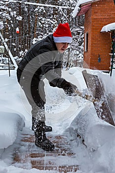 A man cleans steps from snow. In a red hat of Santa Claus and a leather black jacket. In felt boots. Clearing snow from the backya