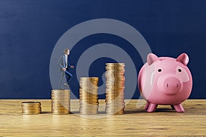 Man climbing stairs to creative pink piggy bank with stacked golden coins on wooden surface and blue wall background. Money