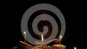 a man in a cloak with a hood is reading a hot book with candles on the floor