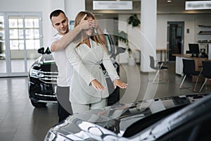 Man close eyes to his wife and makes a surprise buying car. Man and woman in car showroom