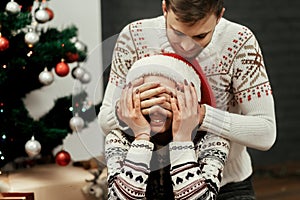 man closing eyes of his happy woman for surprising her with christmas present. cozy family moments in winter holidays at decorate