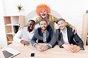 A man in a clown suit is standing next to other men in a bright business office on Fools` Day.