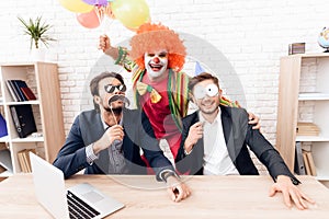 A man in a clown suit is standing next to other men in a bright business office on Fools` Day.