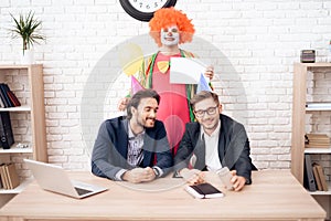 A man in a clown suit is standing next to other men in a bright business office on Fools` Day.