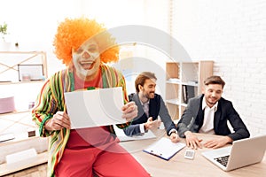 A man in a clown suit stands beside men in business suits, who sit at the desk.