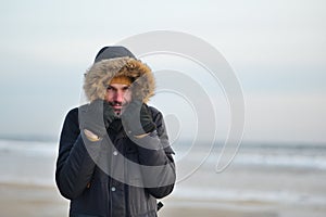 Man in coat holds the hood because of the windy and cold weather at the  sand winter   beach