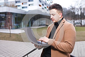 Man in a coat holds a laptop in his hands while standing against the backdrop of a university building. Student working on laptop