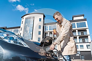 Man with coffee cup in hand inserts plug into the charging socket of electric mobile.