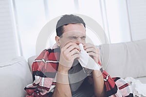 A man with a cold sits on the couch, hiding behind a red rug. He blows his nose into a napkin