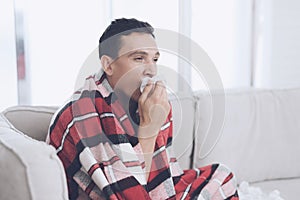 A man with a cold sits on the couch, hiding behind a red rug. He blows his nose into a napkin