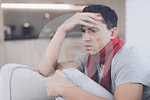 A man with a cold sits on the couch, hiding behind a red rug. He has a headache