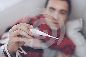 A man with a cold sits on the couch, hiding behind a red rug. He measures the temperature with a digital thermometer