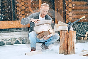 Man collecting chopped firewood on snowy yard for a house fireplace with heavy snowflakes background. Winter countryside holidays