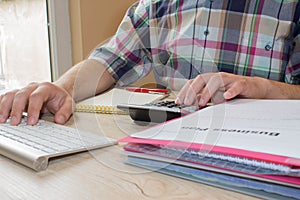 The Man and computer are using a calculator on the table in the office room. The Man sits at the table with a computer and busines