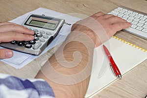 The Man and computer are using a calculator on the table in the office room. The Man sits at the table with a computer and busines