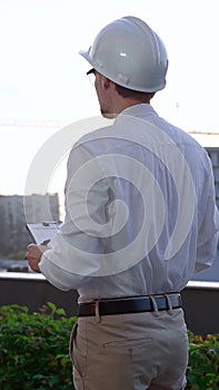 Man a constructive engineer wearing white shirt and hard hat is making notes on a clipboard while inspecting a building