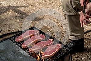 Man cooking beef steaks on a cast iron grlill plate on a camp fire. Campfire cooking. Outdoor BBQ