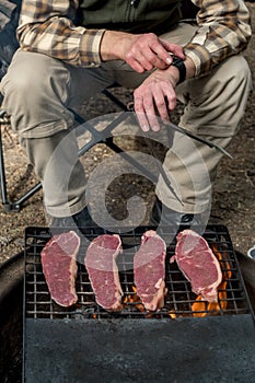 Man cooking beef steaks on a cast iron grlill plate on a camp fire. Campfire cooking. Outdoor BBQ