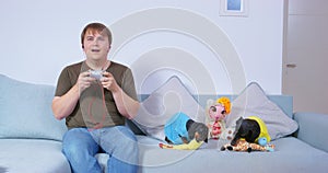 Man on couch holds joystick game console hands, children play dogs soft toys