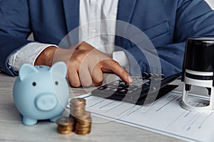 Man is counting profit on a calculator, piggy bank and coins on wooden desk in office. Economy and management financial