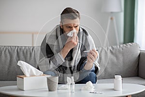 Man covered in blanket sneezing nose and checking for fever