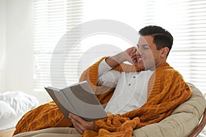 Man covered with warm orange plaid reading book at home