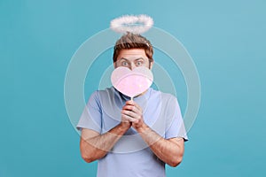 Man covering half of face with pink paper heart on stick, being shy to demonstrate love care.