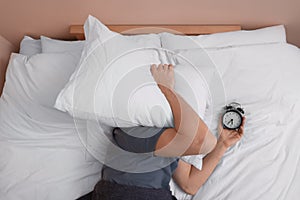 Man covering his face with pillow and holding alarm clock in bed, above view