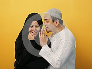 Man covering his mouth and whispering secret to his wife ear, Asian muslim woman looking shocked and excited, opening eyes and