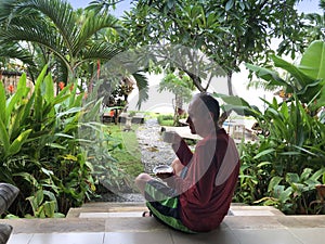 Man with a cup of coffee on the villaâ€™s outdoor terrace has breakfast overlooking the pool and tropical plants. Indonesia, Bali