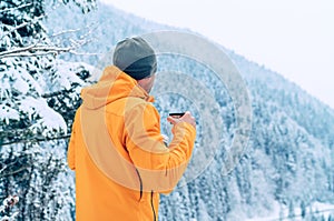 Man with cup drinking a hot drink dressed bright orange softshell jacket and enjoying snowy mountains landscape while he trekking