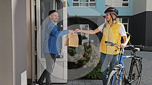 Man customer open door to a food delivery woman wearing yellow thermal backpack on a bike and receives order or package