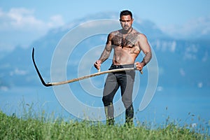 Man cut grass with scythe. Agriculture, farmer working on alps field. Man farmer works on farm, hay making. Sexy farmer