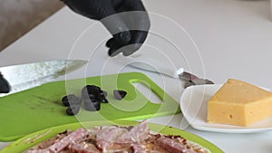 A man cuts olives and places them on a pizza base. Cooking ingredients are laid out nearby. Making pizza at home. Close-up shot
