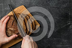 A man cuts a rustic bread with a knife on a cutting board on a dark background. Hands close up