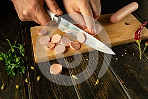 A man cuts vienna sausage on a wooden cutting board. Preparing delicious sandwiches for dinner on the kitchen table at home