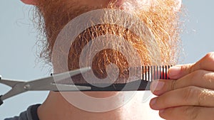 Man cutting his own beard and mustache with scissors and comb. Caucasian red bearded male trimming hair on face at home.