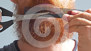 Man cutting his own beard and mustache with scissors and comb. Caucasian red bearded male trimming hair on face at home.