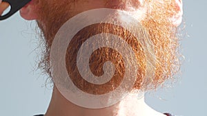 Man cutting his own beard and mustache with scissors and comb. Caucasian red bearded male trimming hair on face at home.