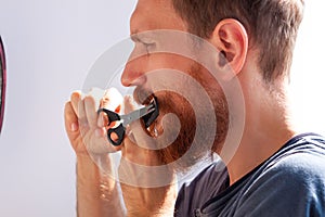 Man cutting his own beard and mustache with scissors and comb. Caucasian red bearded male trimming hair on face at home