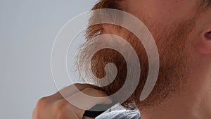 Man cutting his own beard and mustache with scissors and comb. Caucasian red bearded male trimming hair on face at home