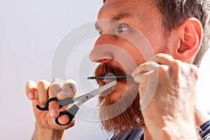 Man cutting his own beard and mustache with scissors and comb. Caucasian red bearded male trimming hair on face at home