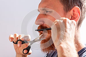 Man cutting his own beard and mustache with scissors and comb. Caucasian red bearded male trimming hair on face at home