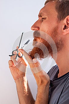 Man cutting his own beard and mustache with scissors and comb. Caucasian red bearded male trimming hair on face at home