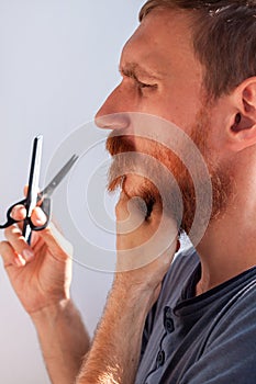 Man cutting his own beard and mustache with scissors and comb. Caucasian red bearded male trimming hair on face at home