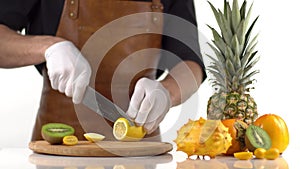 The man is cutting the lemon into slices on the wooden board placed near the pineapple, persimmons and kiwano.