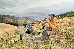 Man cyclist resting after riding electric bike, drinking a cup of tea in the mountains.
